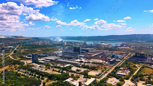 Sprawling industrial site surrounded by green trees and parking lots. Factory buildings and storage areas spread across a wide landscape under a cloudy blue sky.