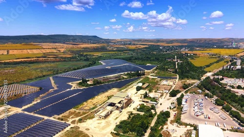 Aerial view of solar panel farm near agricultural fields. Wide shot displays photovoltaic array, green landscape, trucks parked in lot, blue sky with clouds.