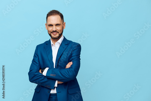 A professional businessman stands with arms crossed against a solid blue background, conveying confidence, poise, clarity, focus, and calm emotion in a clean studio portrait.