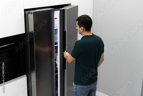 A young man seen from behind opens the door of a large modern side-by-side refrigerator in a bright, minimalist kitchen, looking inside for food. Bachelor's life