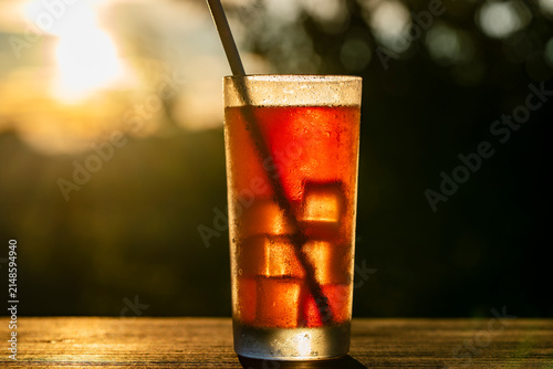 Iced tea drink in glass at golden hour sunset, closeup. Refreshing glass of iced tea or cold brew coffee with ice cubes and straw on wooden table at sunset bokeh background