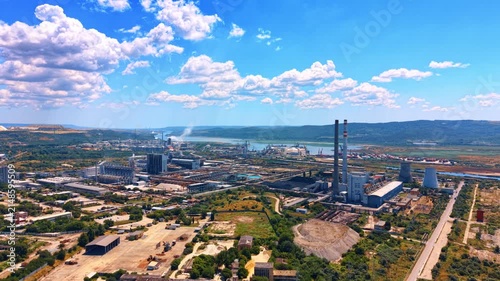 Panoramic aerial view of power station and industrial zone. Tall smokestacks and factory structures stand in a wide industrial valley near water under a bright sky.