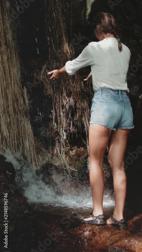 Tourist woman stands near waterfall in tropical jungle forest. Girl picks up water to wash face in hot day. Happy female adult enjoying the water drops. Great wild nature, tourism, travel, holidays