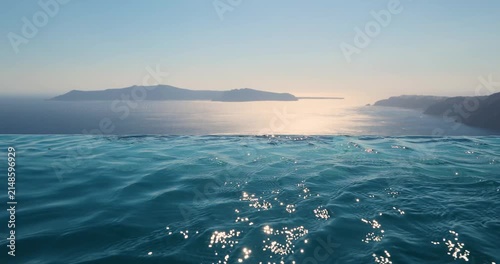 Turquoise blue water ripples in an infinity pool overlooking the caldera of Santorini, Greece as the sunset begins in the background. The wind is blowing. 