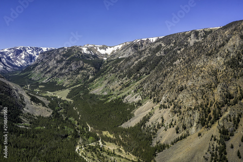 Aerial view of Beartooth Pass showing rugged mountain slopes, evergreen forests, and snow-capped peaks under a clear blue sky in Park County, Wyoming, United States.