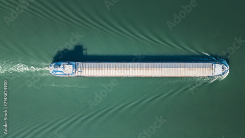 Aerial view of a cargo barge sailing on a river, creating symmetrical wake patterns and a distinct shadow on the water surface in Le Pouzin, Auvergne-Rhone-Alpes, France.