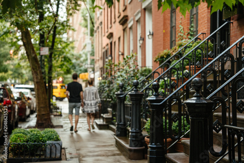 A couple walks along a tree lined New York City brownstone street with red brick buildings and black iron stair railings. Wet sidewalk, parked cars, and greenery create a cozy urban neighborhood