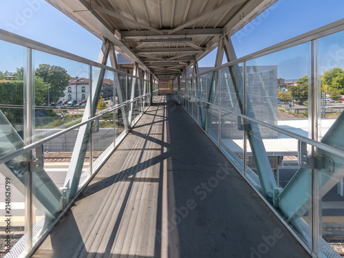 Modern covered pedestrian footbridge over railway tracks at a train station. Interior view of an industrial steel and glass overpass connecting platforms on a bright sunny day.