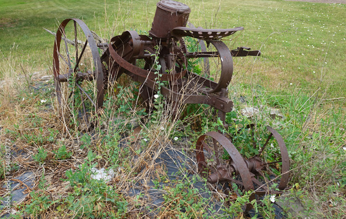 Abandoned horse-drawn antique sickle-bar mower with iron wheels sits overgrown in grass and wildflowers, evoking history and nostalgia for traditional agriculture and past farming equipment