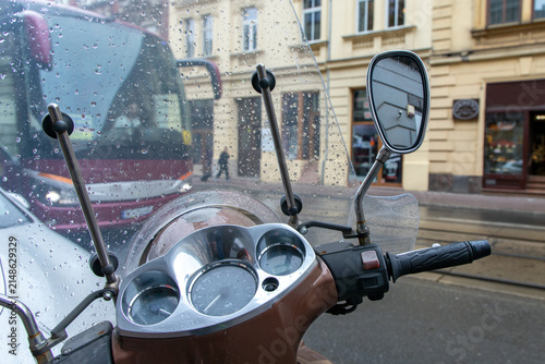 A bus seen through a scooter's windshield in rainy weather. A handlebar-mounted screen made  polycarbonate designed to deflect wind, rain, debris. Chrome mirror, instrument cluster with speedometer. 