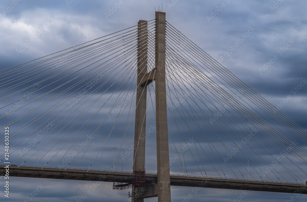 Fototapeta premium Wide view of a large cable-stayed bridge roadway supported by a central concrete tower and symmetrical steel cables under a dramatic, cloudy, blue-grey sky