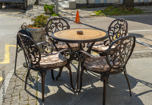 Empty outdoor cafe seating area with ornate wrought iron chairs, padded cushions, and a round wooden table placed on a sunny urban street sidewalk during a bright, warm summer day