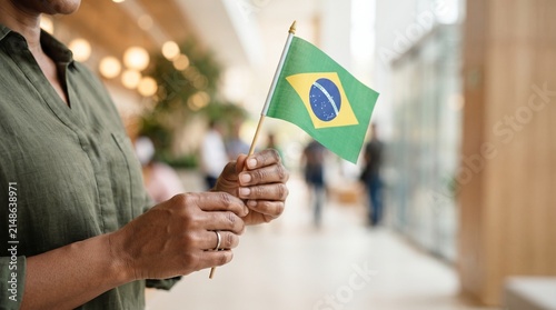 Close-up de mãos segurando pequena bandeira brasileira em ambiente moderno com fundo desfocado, conceito elegante para campanhas da Independência do Brasil