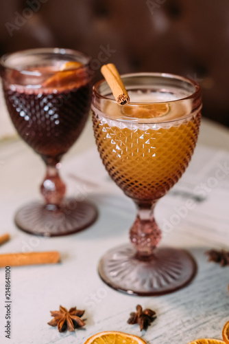 Two decorative glasses filled with orange and dark red seasonal drinks, arranged with cinnamon sticks, star anise and dried citrus slices on a wooden table. Cozy autumn and winter beverage still life 