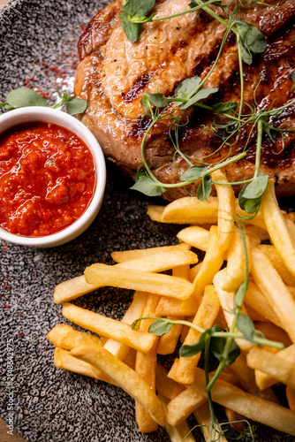 Close-up of a juicy grilled bone-in steak with dark sear marks, served with golden French fries, spicy red dipping sauce and fresh microgreens on a textured ceramic plate.