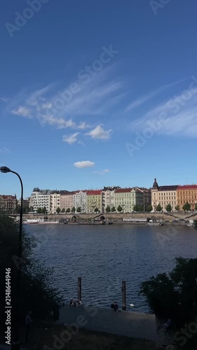 swan glides near riverside promenade while small group of people sit on steps facing wide river, pastel historic buildings lining opposite bank under clear sky river
