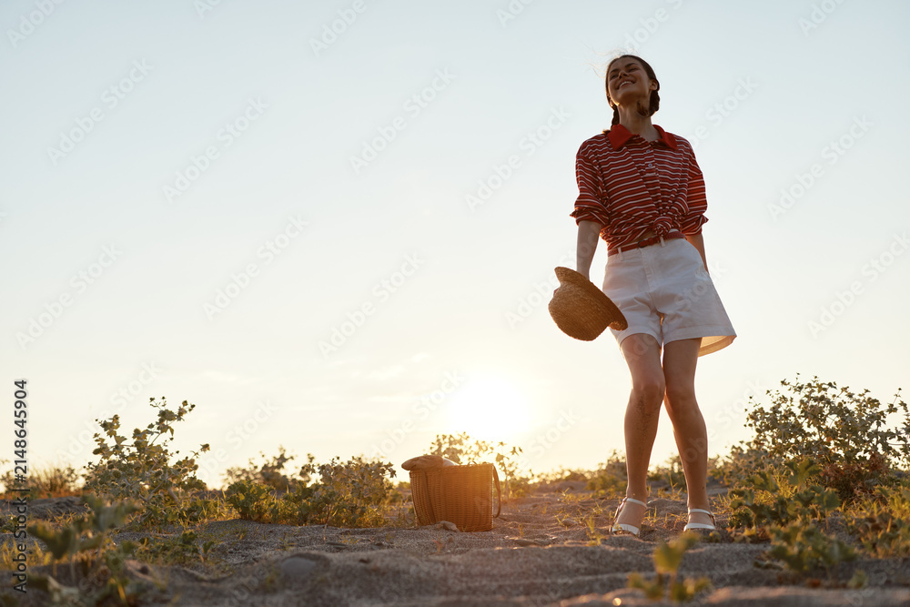 Obraz premium Happy woman enjoying lifestyle during summer picnic on sandy beach at sunset. She holds straw hat and smiles, wearing casual clothes. Concept of freedom, joy, and relaxation.