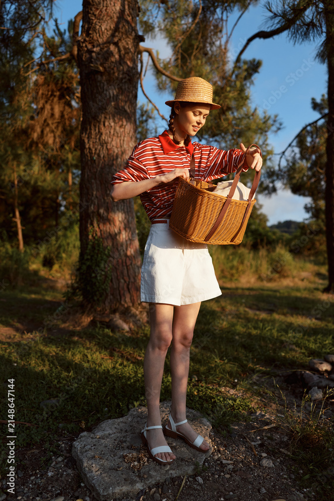 Obraz premium Young woman enjoying a lifestyle picnic outdoors. She holds a woven basket while standing near a tree in the forest. A sunny summer day concept with a beautiful nature setting.
