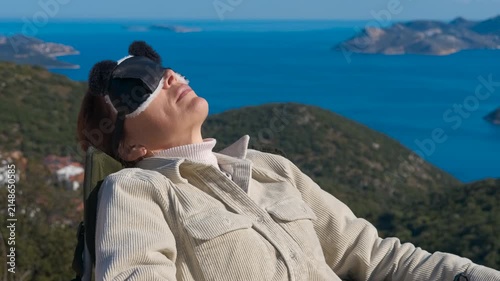 Woman relaxing and sunbathing with beautiful sea view. Content woman taking off a sleep mask and putting on sunglasses while resting in a chair, enjoying a beautiful panoramic view of the sea
