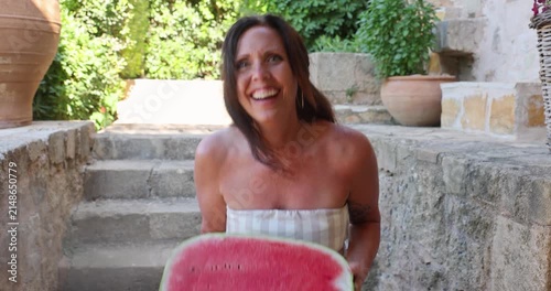 A pretty middle aged woman holding a large, fresh watermelon starts to laugh at the absurd size and silly pose. Her smile is authentic and fun. 