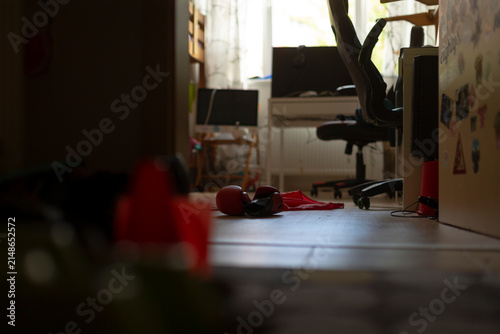 Low, floor-level view into a dim domestic workspace showing a rolling office chair pushed under a desk near a bright window. The foreground is shallow and out