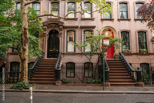 Traditional New York brownstone townhouses with symmetrical staircases, detailed doors, and large windows along a quiet street. Historic brick residential architecture surrounded by trees in a