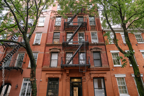 Upward view of a classic New York brownstone apartment building with black metal fire escape and red brick facade framed by green trees. Urban Manhattan architecture featuring detailed windows