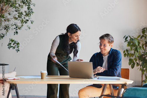 Business professionals collaborating at office desk, discussing project on laptop and documents, sharing ideas during meeting, working together in modern workspace