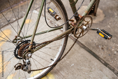 Detailed close up of a bicycle chain, rear gear cassette, and pedals parked on a city sidewalk. Urban transportation concept showing metal components, wheel spokes, and mechanical drivetrain system.