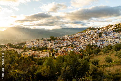 Chefchaouen, Morocco - 14 November 2025: View of the blue-washed buildings of Chefchaouen nestled in the Rif Mountains under a golden sunset sky with olive trees in the foreground.