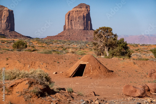 View of a traditional Navajo hogan set against the iconic red sandstone buttes of Monument Valley Tribal Park in the desert landscape of Oljato-Monument Valley, Utah, United States.