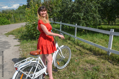 Smiling young woman wearing a red polka dot dress stands beside a white bicycle on a sunny country road, enjoying a relaxed summer moment outdoors among green trees and a wooden fence