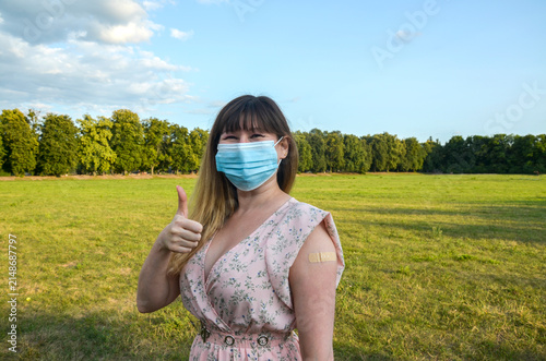 Young woman wearing a surgical mask displays a thumbs up and a small adhesive bandage on her arm after receiving a vaccine in a sunny park, conveying health, relief and community protection