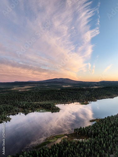 Aerial view of Perilajarvi lake surrounded by dense coniferous forests and distant hills under a dramatic sunset sky with wispy clouds in Enontekio, Lappi, Finland.