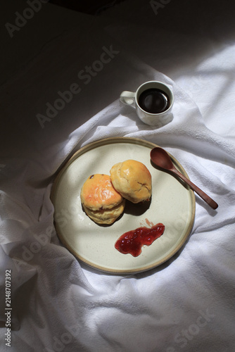Scones with dollop of jam beside steaming cup of coffee, bathed in dappled sunlight. Freshly baked in morning.