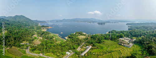 Aerial view of Wadaslintang Reservoir surrounded by lush green hills, terraced rice fields, and a winding road under a clear sky in Wadaslintang, Central Java, Indonesia.