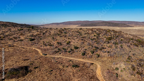Aerial view of a winding dirt road traversing the arid, hilly savanna landscape under a clear blue sky in Modimolle, Limpopo, South Africa.