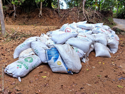 Stacked white and blue feed sacks on rural red dirt ground by lush forest vegetation