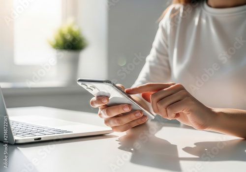 Close up shot of young professional woman sitting at desk using modern smartphone device near laptop with blurred office interior background view.