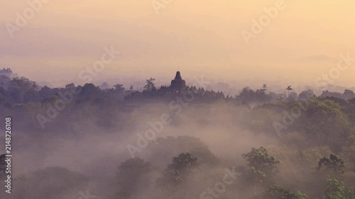Aerial view of Borobudur Temple surrounded by lush trees and morning mist during sunrise in Kecamatan Borobudur, Jawa Tengah, Indonesia.