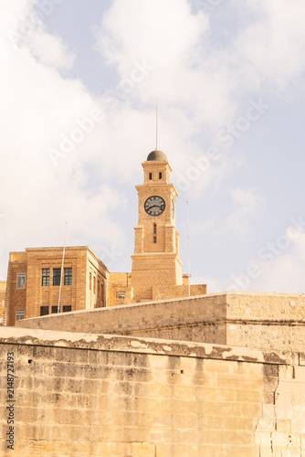 Valletta, Malta - 15 July 2022: View of the historic clock tower of the former Garrison Chapel in Valletta, featuring golden limestone architecture against a soft blue sky.