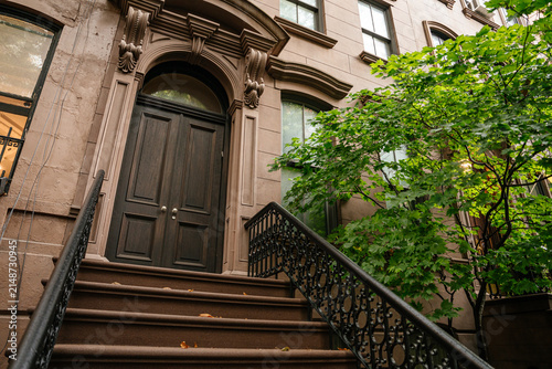 Elegant New York brownstone entrance featuring detailed stone arch, dark wooden door, and decorative iron railing. A leafy tree adds greenery to the classic urban residential facade.