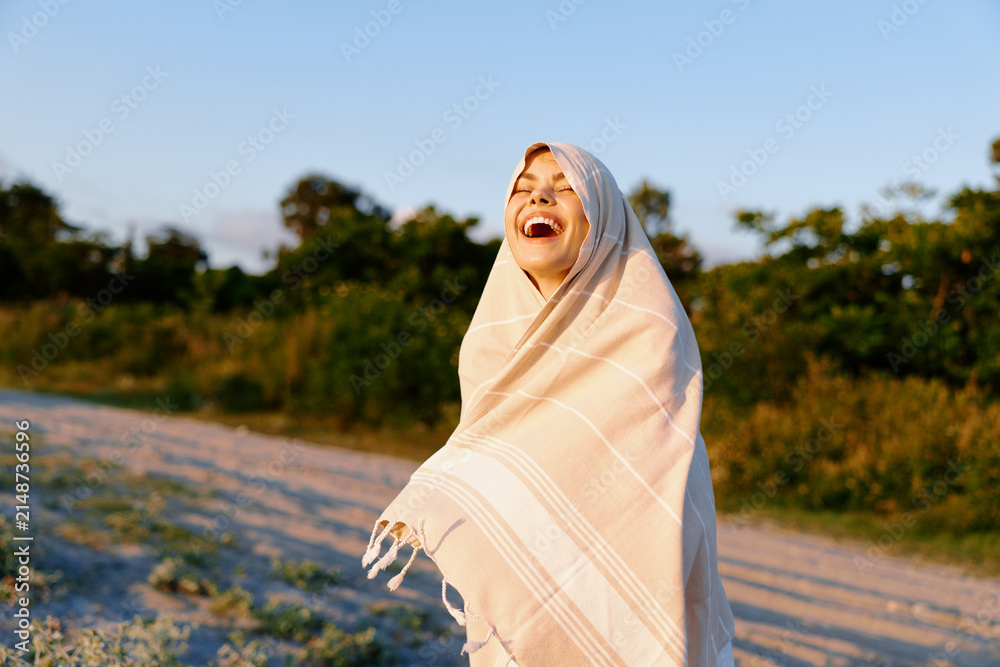Obraz premium Young woman wrapped in a light blanket laughs outdoors on a sunny beach path, radiating joy, wellness, and carefree summer vibes.