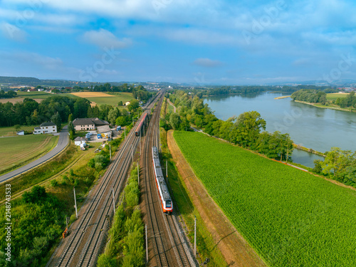 Aerial view of local trains traveling along the Danube river through green fields and rural landscape in Sarling, Lower Austria, Austria.
