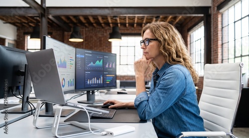Focused professional woman in glasses analyzing financial data on multiple computer screens in a modern office setting