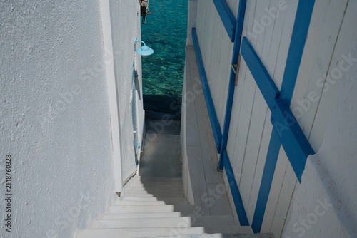 Foto A narrow passage between whitewashed Greek buildings reveals vivid turquoise water beyond, combining blue accents, traditional island architecture and the bright colors of the Aegean