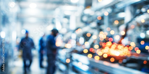 Blurred image of industrial workers in a manufacturing facility, showcasing machinery and production processes. Lights create a bokeh effect in the background.