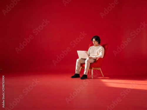 Young Asian Man Focus Using Laptop in Minimalist Modern Armchair with Copy Space of Red Studio Background