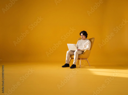 Young Asian Man Focus Using Laptop in Minimalist Modern Armchair with Copy Space of Yellow Studio Background