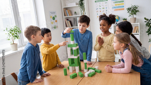 Group of diverse elementary students collaborates on building a tall structure using green and white wooden blocks on a large classroom table.